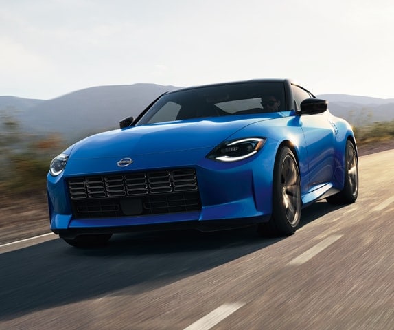 Angled front view of a blue 2024 Nissan Z driving fast on a road lined with brush