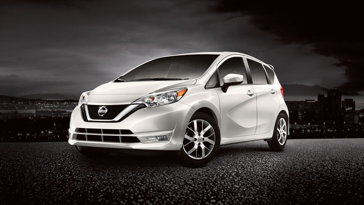 A white Nissan Versa Note parked on asphalt with a city in the background