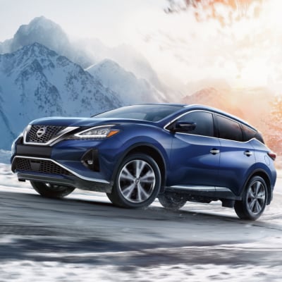 A blue 2024 Nissan Murano driving up a snow covered road with a mountain and trees in the background